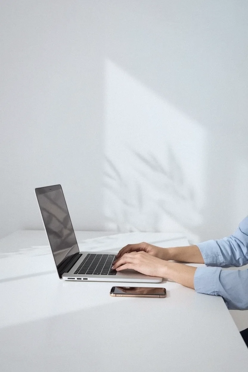 Mãos digitando em um notebook sobre uma mesa branca, com a luz da janela na parede